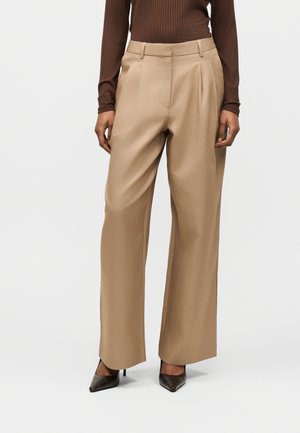 Femme portant un pantalon beige taille haute à jambes larges, un haut marron côtelé à manches longues et des talons noirs à bouts pointus, debout devant un fond uni.