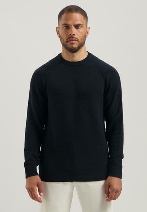 Man met kort haar en een baard draagt een eenvoudige zwarte sweater met lange mouwen en lichtgekleurde broek tegen een neutrale achtergrond.