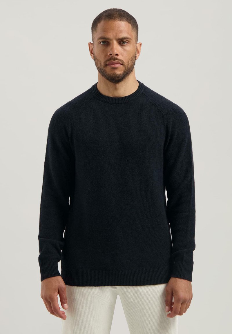 Mann mit kurzem Haar und Bart trägt einen schlichten schwarzen Pullover mit langen Ärmeln und helle Hose vor einem neutralen Hintergrund.