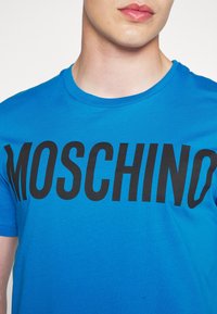 Osoba oblečená v jasně modrém tričku s velkým černým textem "MOSCHINO" napříč hrudníkem, viditelným od krku až po střed trupu.