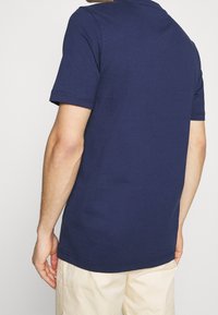 Námořnická modrá bavlněná tričko s krátkými rukávy, kulatým výstřihem a šitými okraji. Zobrazeno zezadu, ukazující jednoduchý design.