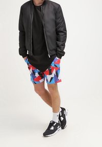 Chaqueta bomber negra, camiseta negra, bañador geométrico colorido con patrones rojos, azules y blancos, zapatos deportivos negros con acentos blancos.