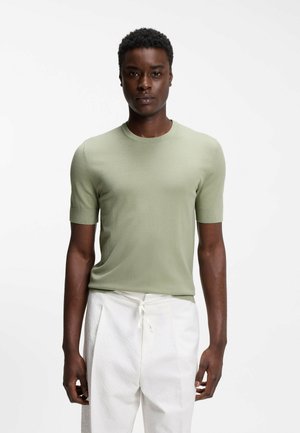 Młody mężczyzna stoi, ubrany w jasnozieloną dzianinową koszulkę z krótkim rękawem i białe spodnie z wiązanym paskiem, na jednolitym białym tle.