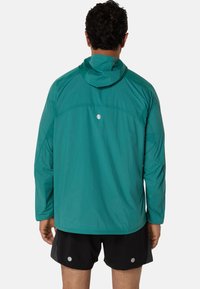 Veste légère à capuche teal avec des manches longues, présentant un accent réfléchissant à l'arrière et un tissu respirant, associée à un short noir.