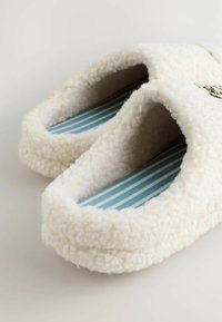 Vita, fluffiga tofflor med en plyschliknande textur, som har en blå och vit randig sula. Rundad design med öppen bakdel för enkel påtagning.