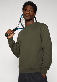 Bărbat îmbrăcat într-un hanorac verde măsliniu, ținând o rachetă de tenis neagră și albastră pe umăr, pe un fundal simplu.