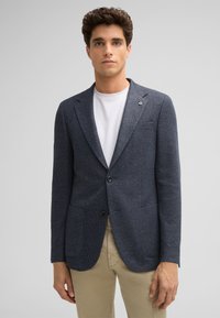 Navy-gestreifter Blazer mit zwei vorderen Taschen, Ein-Knopf-Verschluss und Reversdetails, kombiniert mit einem weißen T-Shirt und beigefarbenen Hosenträgern.