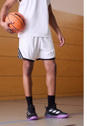Persoon in witte basketbalshorts en shirt houdt een oranje basketbal vast, staat op een binnenveld en draagt zwarte en paarse sportschoenen en zwarte sokken.