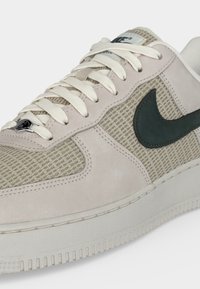 Μπεζ και γκρι αθλητικό παπούτσι Nike με υφή πλέγματος, λευκά κορδόνια και μαύρο λογότυπο Swoosh στο πλάι, εμφανιζόμενο από εμπρόσθια πλευρική γωνία.