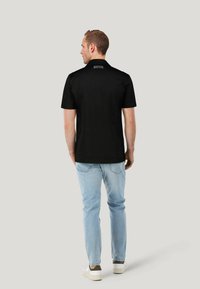 Czarna koszulka polo z krótkim rękawem i płaskim kołnierzykiem, noszona z jasnoniebieskimi dżinsami i białymi sneakersami, widoczna z tyłu.