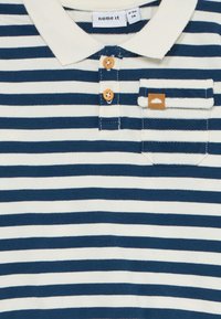 Camisa polo a rayas blanca y azul marino con cuello crema, tapeta de dos botones de madera y pequeño bolsillo en el pecho a rayas con detalle de cuero.