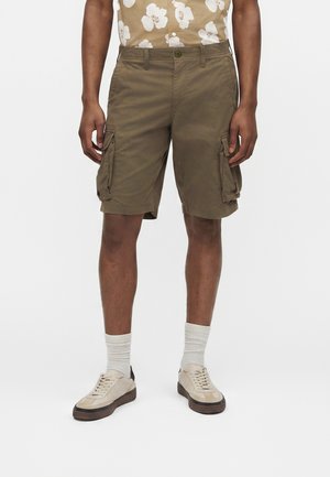 Mies pukeutunut ruskeisiin cargo-shortseihin, beige kukkakuosinen lyhythihainen paita pukeutuneena housuihin, valkoiset tähtisukat ja beiget lenkkarit ruskeilla pohjilla.