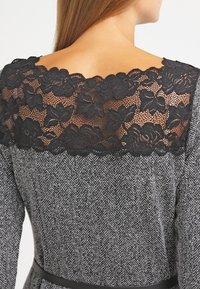 Abito grigio con un motivo a spina di pesce testurizzato, con una scollatura in pizzo nero e un design floreale, abbinato a una cintura nera in vita.