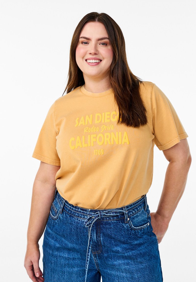 Camiseta de algodón color mostaza con el texto "San Diego Rodeo Drive California 1769" en negrita, combinada con jeans de mezclilla azul de cintura alta.