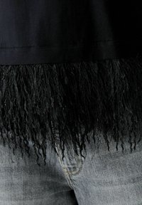 Schwarzes Oberteil mit Fransen am Saum, texturiert mit langen schwarzen Strähnen. Kombiniert mit hellgrauen Jeans, die eine sichtbare Naht aufweisen.