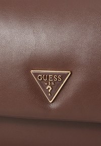 Brun læroverflate med et gulltrimmet trekantet Guess USA-logo med et spørsmålstegn i midten.