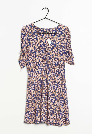 Niebieska sukienka z białym i pomarańczowym wzorem stokrotek, krótkie rękawy z wiązaniami, dekolt w kształcie litery V z detalem wiązania z przodu, zawieszona na czarnym wieszaku.