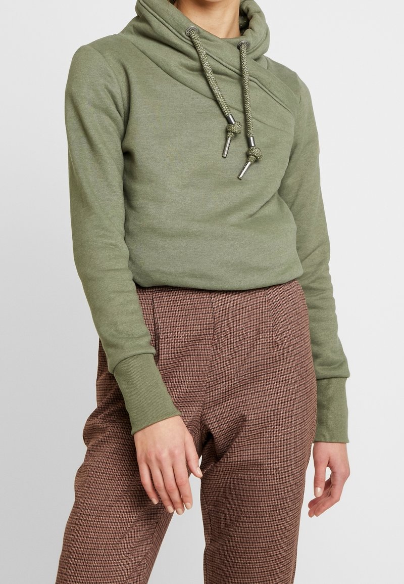 Donna che indossa un felpa verde oliva con stringhe e pantaloni a quadri marroni, in piedi di fronte a uno sfondo semplice.