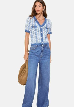 Frau trägt eine blau-weiß karierte Kurzarm-Strickjacke mit Knöpfen, hoch taillierte, weit geschnittene Jeans und hält eine beige Wildledertasche.