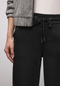 Schwarze Jogginghosen mit Kordelzug an der Taille, seitlichen Taschen und einem glatten, strukturierten Stoff. Darüber ist eine graue, strukturierte Jacke mit Reißverschluss sichtbar.