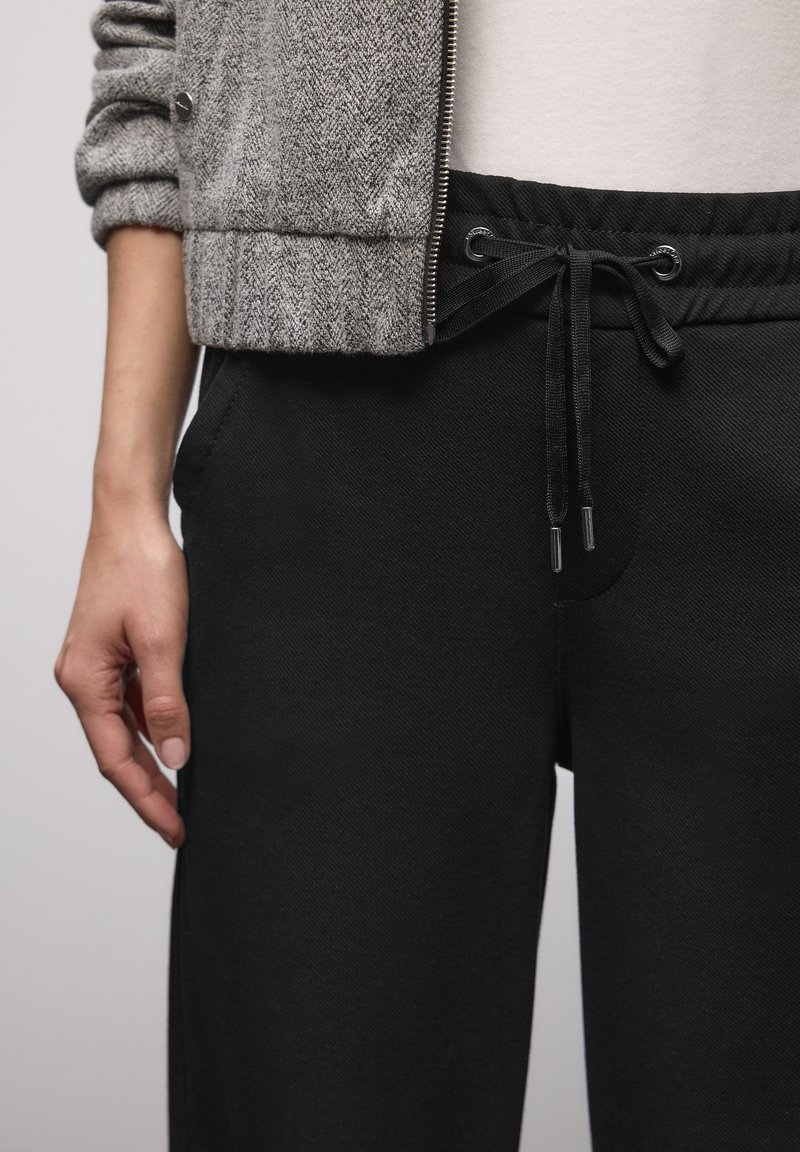 Schwarze Jogginghosen mit Kordelzug an der Taille, seitlichen Taschen und einem glatten, strukturierten Stoff. Darüber ist eine graue, strukturierte Jacke mit Reißverschluss sichtbar.