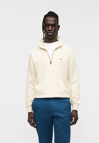 Κρεμ φούτερ με φερμουάρ μέχρι τη μέση και μπροστινή τσέπη, με μικρό λογότυπο, σε συνδυασμό με μπλε παντελόνι. Το ύφασμα φαίνεται λείο και ελαφρύ.