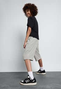 T-shirt en coton noir, shorts gris arrivant aux genoux avec logo, chaussettes blanches, baskets noir et blanc, cheveux bouclés, avec un fond neutre.
