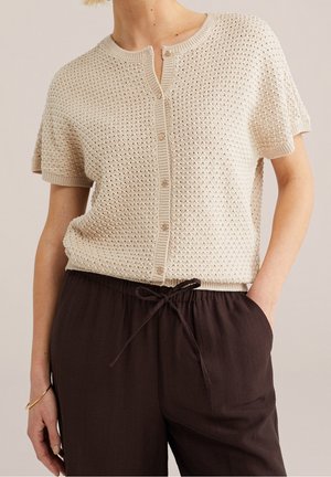 Personne portant un haut beige en maille à manches courtes boutonné et un pantalon marron foncé à cordon, une main dans la poche et un bracelet au poignet.