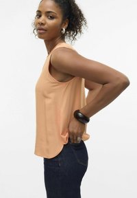 Top senza maniche color pesca con orlo arrotondato e texture liscia, abbinato a jeans in denim scuro. Presenta un grande bracciale in legno su un polso.