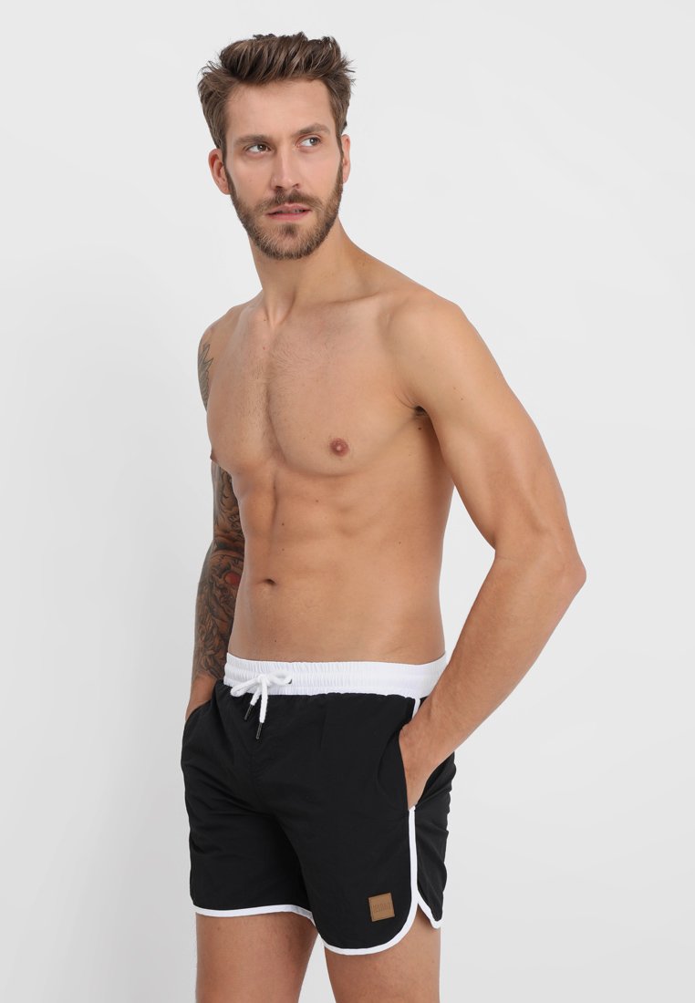 Man zonder hemd met baard en tatoeage op de linkerarm, draagt zwarte zwemshorts met witte tailleband en rand, staat met handen in de zakken, kijkt naar rechts.