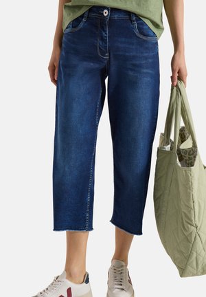 Personne portant un jean bleu foncé raccourci avec des ourlets effilochés, des baskets blanches et tenant un sac cabas matelassé vert clair.