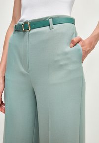 Jasnozielone spodnie typu szerokiego z g�ładką fakturą, z bocznymi kieszeniami i dopasowanym niebiesko-zielonym paskiem z detalem w postaci złotego zapięcia.