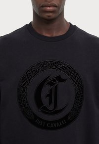 Felpa nera con un grande logo circolare in rilievo che presenta una "C" stilizzata e un design di serpente intrecciato, realizzata in tessuto morbido.