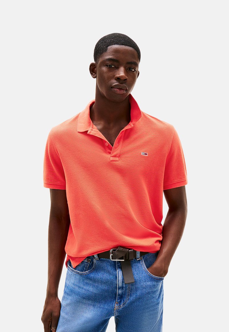 Polo de manga corta, de color coral brillante, con un acabado texturizado, que presenta un cuello y un pequeño logo. Combinado con vaqueros azules y un cinturón negro.