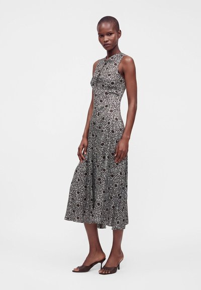Femme debout portant une robe midi fleurie noire et blanche sans manches avec des mules marron ouvertes à talons sur fond blanc.