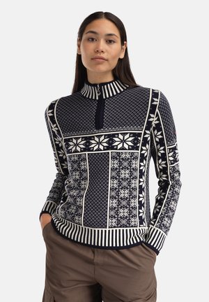 Kvinne iført en marineblå og hvit genser med glidelås i halsen, dekorert med geometriske mønstre og snøfnugg, kombinert med brune bukser, hendene i lommene.