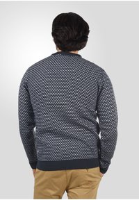 Mørkeblå sweater med et geometrisk hvidt mønster, ribbede manchetter og kant, samt rund halsudskæring. Stoffet ser tekstureret og strikket ud.