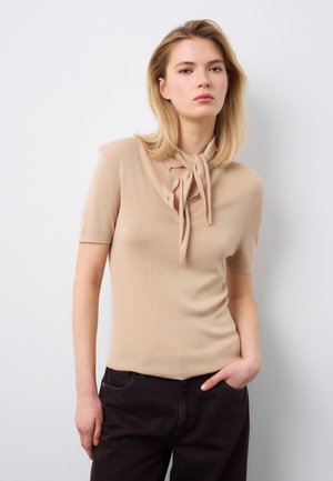 Giovane donna con capelli biondi indossa una maglietta beige a maniche corte con scollo annodato e pantaloni neri, in piedi contro una parete bianca semplice.