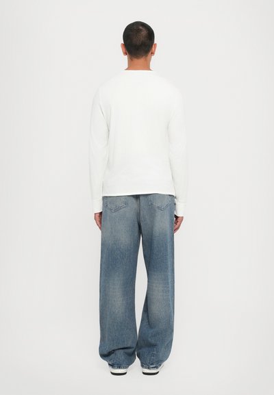 Chemise blanche à manches longues associée à un jean en denim bleu clair à coupe ample, avec une légère décoloration et deux poches arrière ornées de coutures.