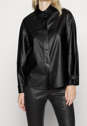 Kvinde iført en sort skjortejakke i imiteret læder med lange ærmer og matchende sorte bukser i imiteret læder.