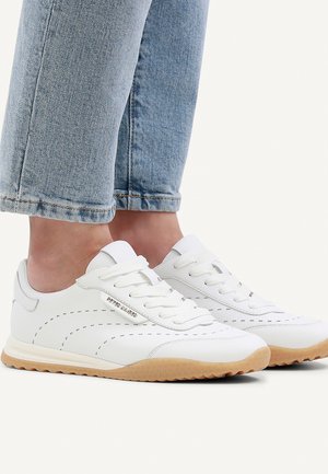 White leather sneakers with gum soles worn with cuffed light blue denim jeans, showing stitched detailing and branded metal tag on sides.