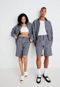 Gestreepte stoffen set in marineblauw en wit, met een knoopjasje, shorts en een cropped top. Schoenen: witte slides en zwarte instappers.
