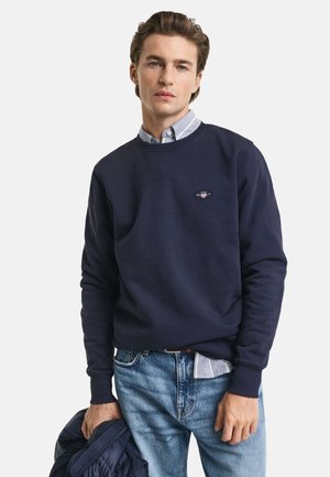 Navyblauwe sweatshirt met ronde hals, geribbelde manchetten en geborduurd logo op de borst, gedragen over een lichtblauwe overhemd met kraag.