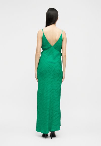 Γυναίκα με μακριά σκουρόχρωμα μαλλιά, που φοράει ένα πράσινο μακρύ φόρεμα χωρίς μανίκια με βαθύ V στην πλάτη και διακριτικό σχέδιο, στέκεται με τα χέρια χαλαρά στο πλευρό.