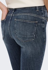 Tamnoplave traperice uskog kroja, s visokim strukom i stražnjim džepovima s kontrastnim šavovima. Glatka tekstura i minimalno blijeđenje boje.