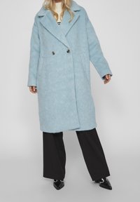 Cappotto oversized in misto lana di colore azzurro chiaro con una texture morbida, colletto a scollo a V, chiusura doppiopetto e tasche laterali. Indossato sopra pantaloni neri.