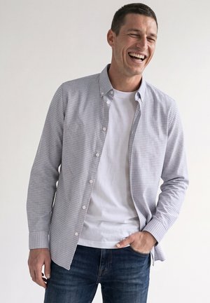 Mann lacht mit geschlossenen Augen, trägt ein weißes T-Shirt, ein aufgeknöpftes Hemd mit kleinem Karo und blaue Jeans vor einem einfarbigen Hintergrund.