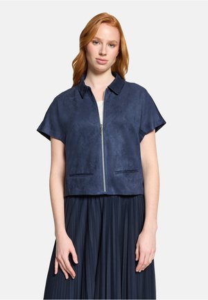 Femme portant une veste à manches courtes bleu marine avec fermeture éclair et col, ainsi qu'une jupe plissée bleu marine, debout devant un fond uni.