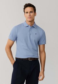 Mężczyzna z ręką w kieszeni, ubrany w jasnoniebieską koszulkę polo i ciemne spodnie, stojący na tle gładkiego, jasnoszarego tła.