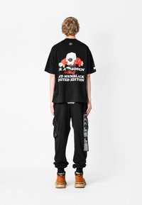 Camiseta negra de gran tamaño con estampado gráfico en la parte posterior, combinada con pantalones cargo negros que tienen un bolsillo lateral y acentos de texto blanco. Calzado marrón.